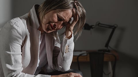 A woman in a white blazer sits with her head in her hand, showing distress.