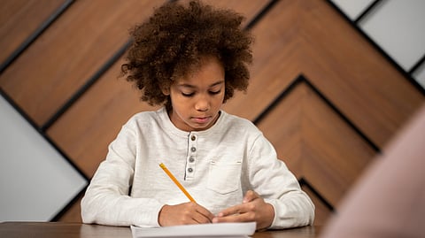 Child with curly hair concentrating on writing with a pencil at a wooden desk.