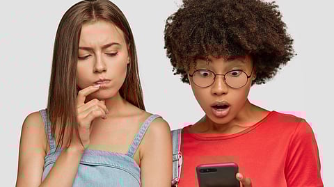 Two young women react to something on a smartphone, the one on the left looks puzzled.