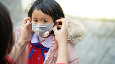A young child wearing a grey face mask is being helped by an adult.