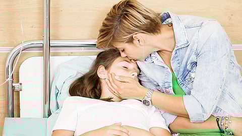 A woman gently kisses a girl's forehead as she rests in a hospital bed.
