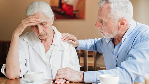 An elderly man comforts a distressed elderly woman in a cafe, gently touching her shoulder. 