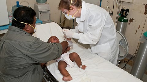 A healthcare worker wearing protective gear administers an injection to a baby crying on a hospital bed.