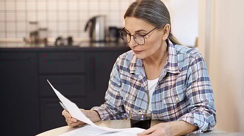 An older woman in glasses reads papers intently at a kitchen table. 