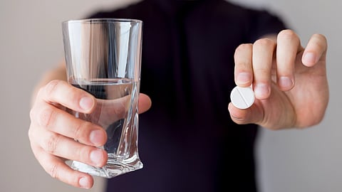 A person holds a glass of water in the left hand and a round white pill in the right hand.
