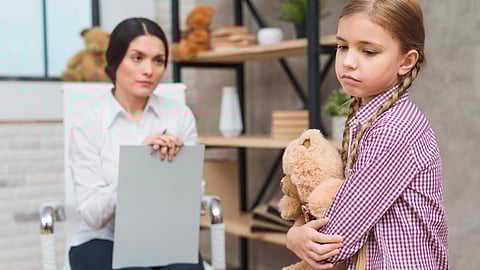A young girl with a concerned expression hugs a teddy bear,a woman attentively listens, sitting near a shelf with toys.
