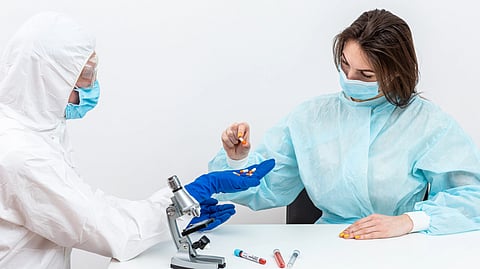 Two scientists in protective gear and masks analyze pills in a lab.