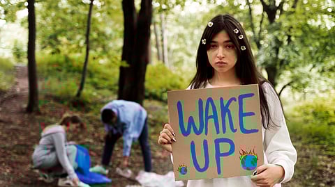 Young woman in forest holds "Wake Up" sign with burning Earth illustrations, looking serious.