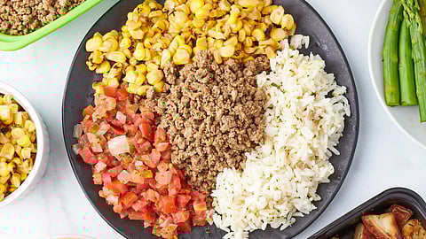 Overhead flat-lay of a portioned meal bowl with ground meat, rice, corn, and pico de gallo, surrounded by prepped food containers — illustrating routine, balanced eating.