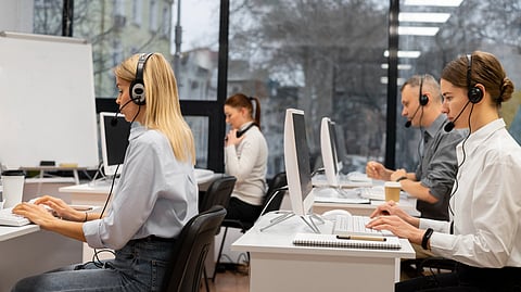 A group of call center agents sit at desks in a modern office, wearing headsets and working on computers.