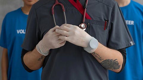 A doctor in grey scrubs with marron stethoscope around neck and 2 people standing behind him in blue tees.