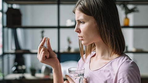 Woman in a purple shirt intently examines a pill while holding a glass of water.