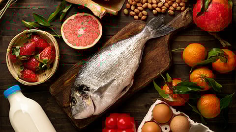 A wooden table with fresh ingredients: whole fish on a board, strawberries, grapefruit, eggs, milk, chicken, almonds, bell peppers, pomegranate, tangerines.