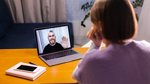 A teen age girl sits at a table, engaged in a video call on a laptop.