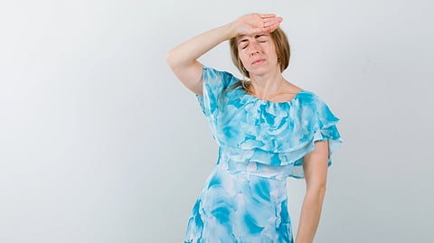 A woman in a blue floral dress stands with her eyes closed, hand on forehead.