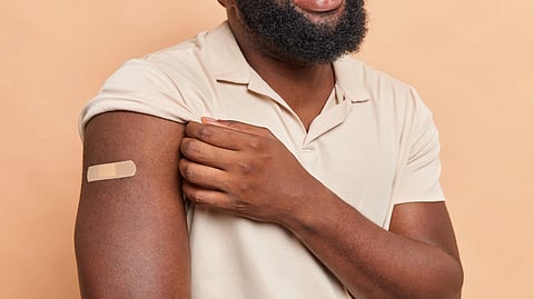 A smiling person with a beard, wearing a beige shirt, shows off a bandage on their upper arm.