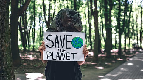 A person stands in a forest wearing a black plastic bag over their head, holding a "Save the Planet" sign with a drawing of Earth.
