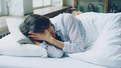 A woman in a striped shirt lies in bed, face buried in her hands, conveying sadness or exhaustion.