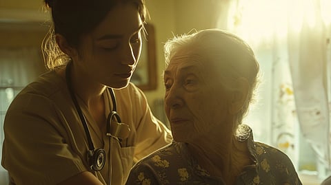 A caregiver with a stethoscope gently supports an elderly woman in a sunlit room.