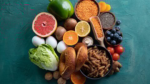A variety of fresh foods on a teal surface, including fruits, vegetables, grains, and seeds.