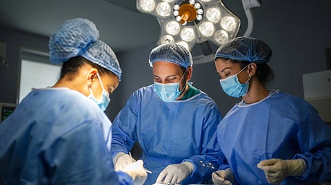 Three surgeons in blue scrubs and masks focus intently on a procedure under bright operating room light.