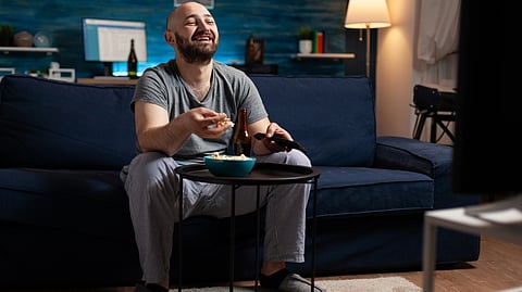 A man in pajamas laughs while watching TV, sitting on a blue couch in a cozy living room.