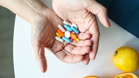 Hands holding colorful capsules and tablets above a white table with two oranges nearby.