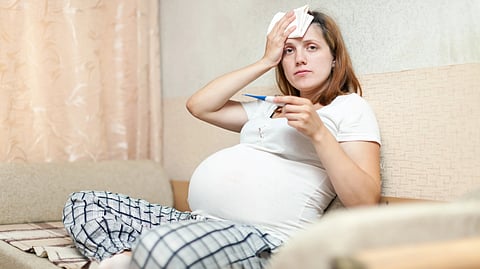 Pregnant woman in pajamas looks unwell, sitting on a couch holding a thermometer and pressing a tissue to her forehead.