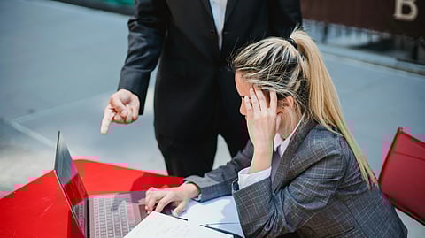 A woman in a suit sits at a red table, looking stressed as she works on a laptop. 