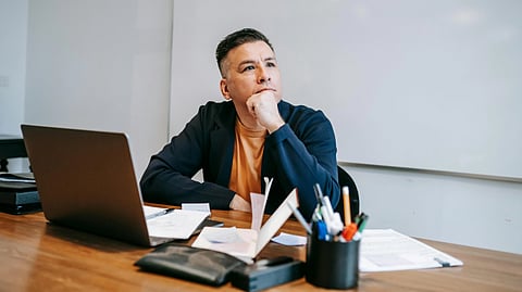 A person sits at a table with a laptop, papers, and pens, resting their chin on their hand, looking thoughtful. 