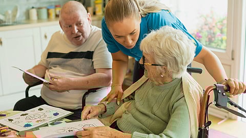 A caregiver in a blue uniform leans over to assist an elderly woman in a wheelchair with coloring.