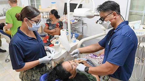 Dentists treat patients in a clinic. One dentist works on a patient lying in a chair, while an assistant observes. 