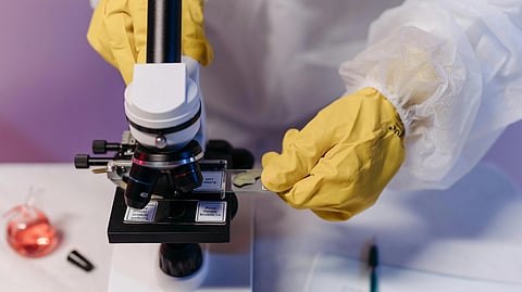 Scientist in protective gear adjusts a slide under a microscope, with yellow gloves. 