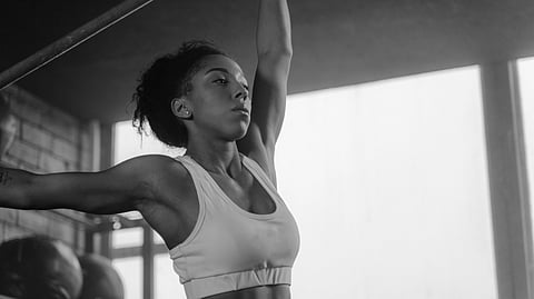 A woman in a gym, wearing a sports bra, stretches with one arm raised, looking focused.