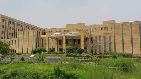A large, modern AIIMS building with a light brown exterior, tall windows, and greenery all around.