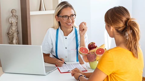 A nutritionist in a white coat, with a blue measuring tape, talks to a client at a desk, fresh fruit in a bowl and a laptop.