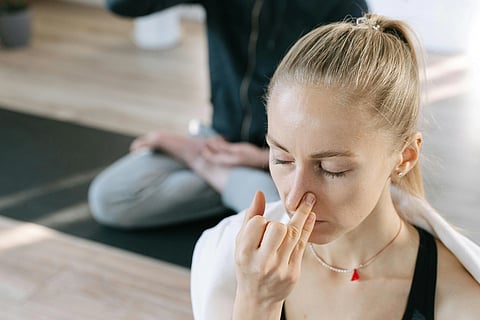 Woman practising pranayama, alternate nostril breathing performed seated indoors with a second person visible in the background, suggesting supervised practice