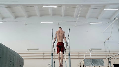 A shirtless athlete in red shorts practices on parallel bars in a spacious gymnasium.