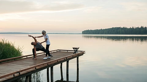 Two people do yoga on a wooden dock at sunrise, one helping the other stretch.