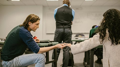A male student in a classroom discreetly hands a paper to a female student. 