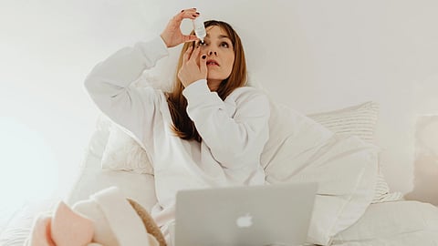 Woman in a white sweater sitting on a bed using eye drops.