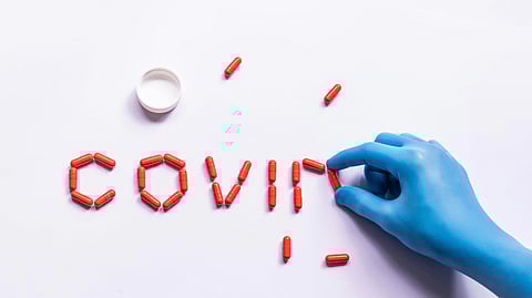 A blue-gloved hand arranges red capsules to form the word "COVID" on a white background.