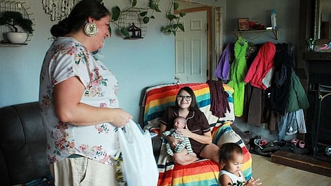 A woman stands smiling with a bag, facing a mother seated on a rainbow-striped chair holding a baby. 