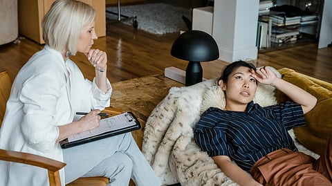 A therapist with a clipboard attentively listens to a woman lying on a couch.