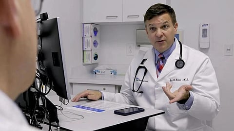 Doctor in white coat talks to a patient at a desk.