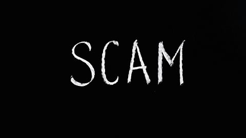 The image features the word "SCAM" written in bold, white chalk-like letters against a solid black background.