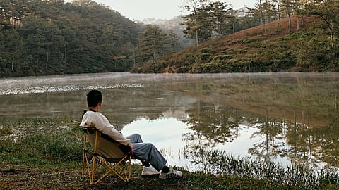 A person sits in a chair by a tranquil lake, surrounded by mist and pine trees, under a clear sky. 