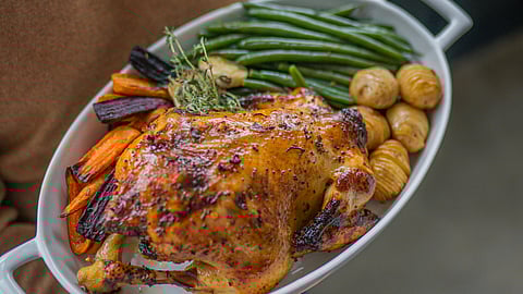 Person in a brown sweater holds a white dish with roasted chicken, colorful carrots, green beans, and potatoes.