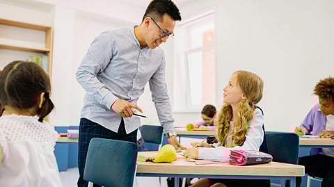 A teacher smiles and helps a young girl at her desk in a classroom.