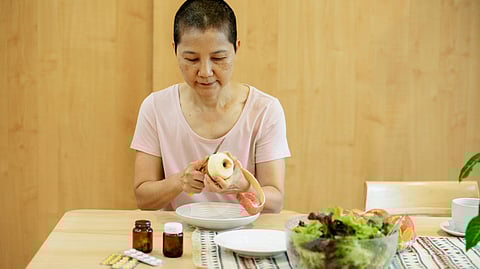A woman with short hair, wearing a pink shirt, peels an apple at a wooden table. 
